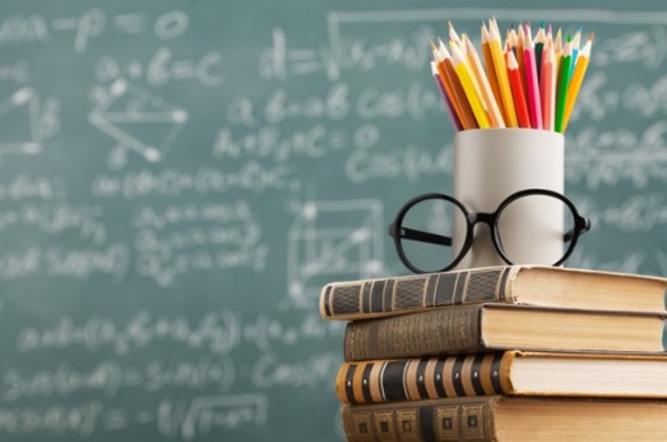 A pair of glasses and a container of colored pencils sits atop some old leather bound books. In the background is a chalkboard filled with math concepts.