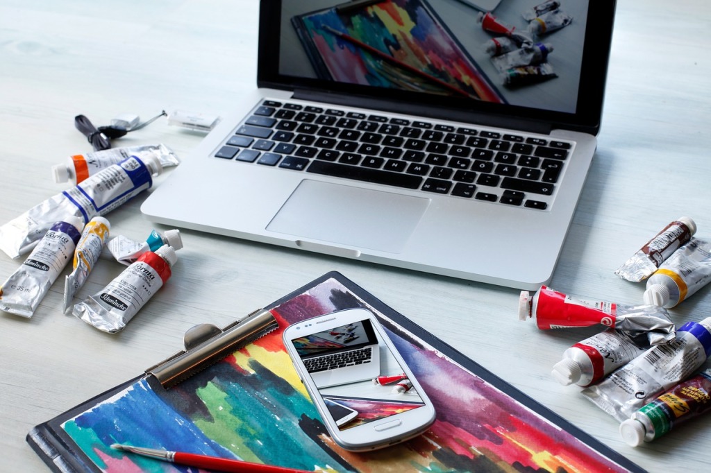 An open laptop sits on a desk with colorful tubes of paint.